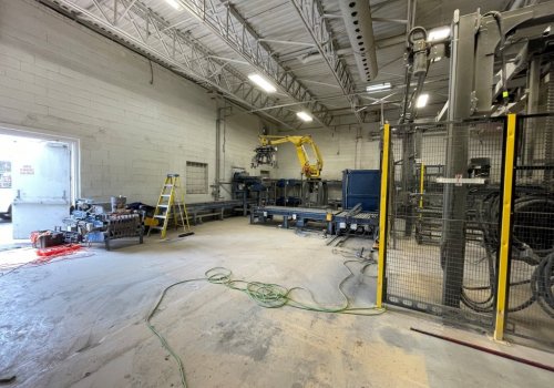 Zone industrielle intérieure avec un bras robotisé jaune, des équipements de manutention, des convoyeurs et divers outils au sol dans un espace en cours d’aménagement.