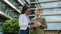 Deux personnes discutent ensemble à l’extérieur d’un immeuble moderne en consultant une tablette. 