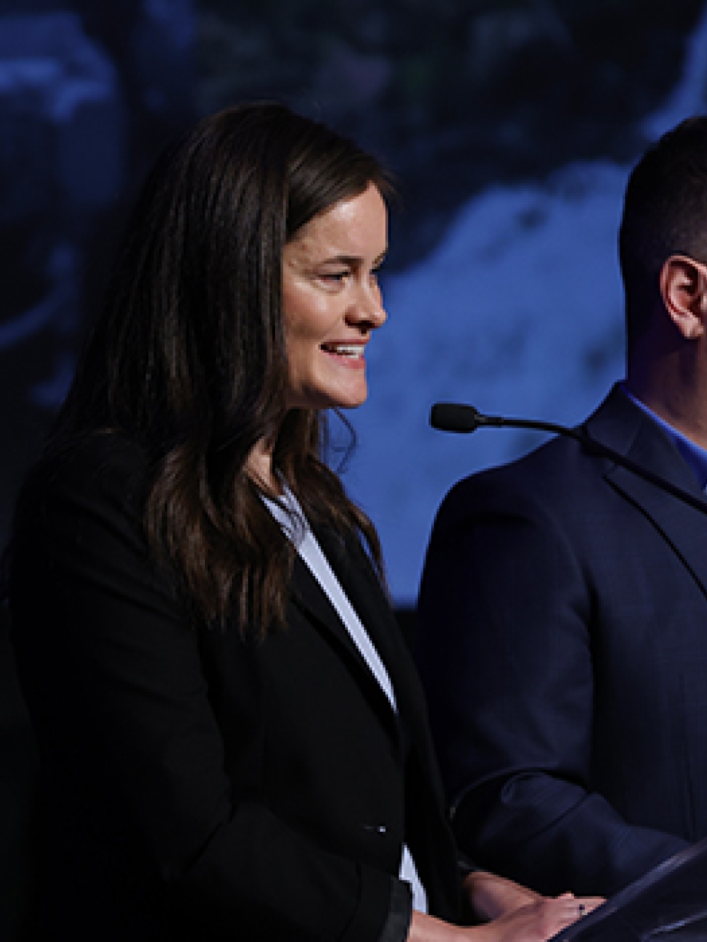 Deux personnes en tenue professionnelle debout derrière un lutrin, parlant devant des micros. 