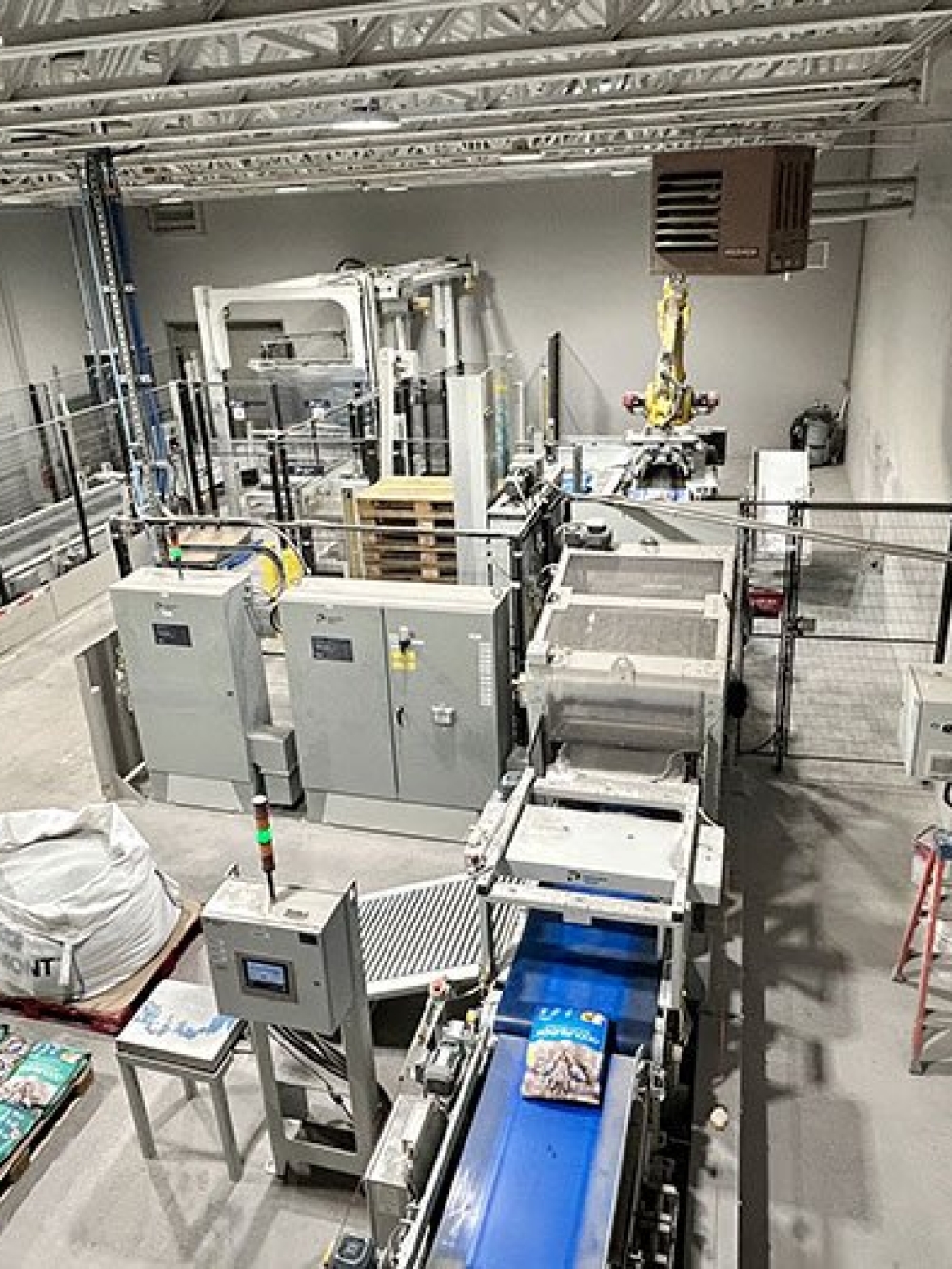 Vue d’ensemble d’une installation industrielle automatisée avec convoyeurs, postes d’emballage, équipements de manutention et sacs de produits, dans un entrepôt portant le logo Intersand sur le mur.