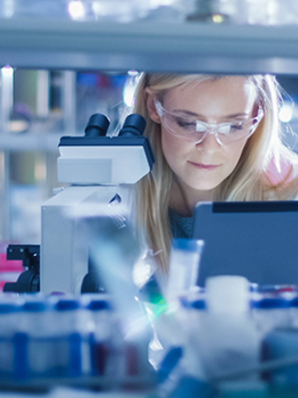 Personne en laboratoire travaillant avec un microscope, entourée de matériel scientifique et de flacons.
