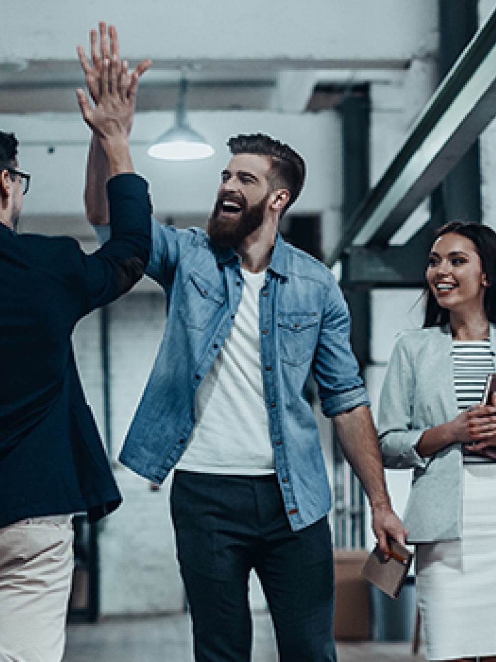 Groupe de collègues dans un bureau industriel, dont deux se donnent un high‑five tandis qu’une autre personne tient des documents et qu’une quatrième est assise à l’arrière avec un ordinateur portable.