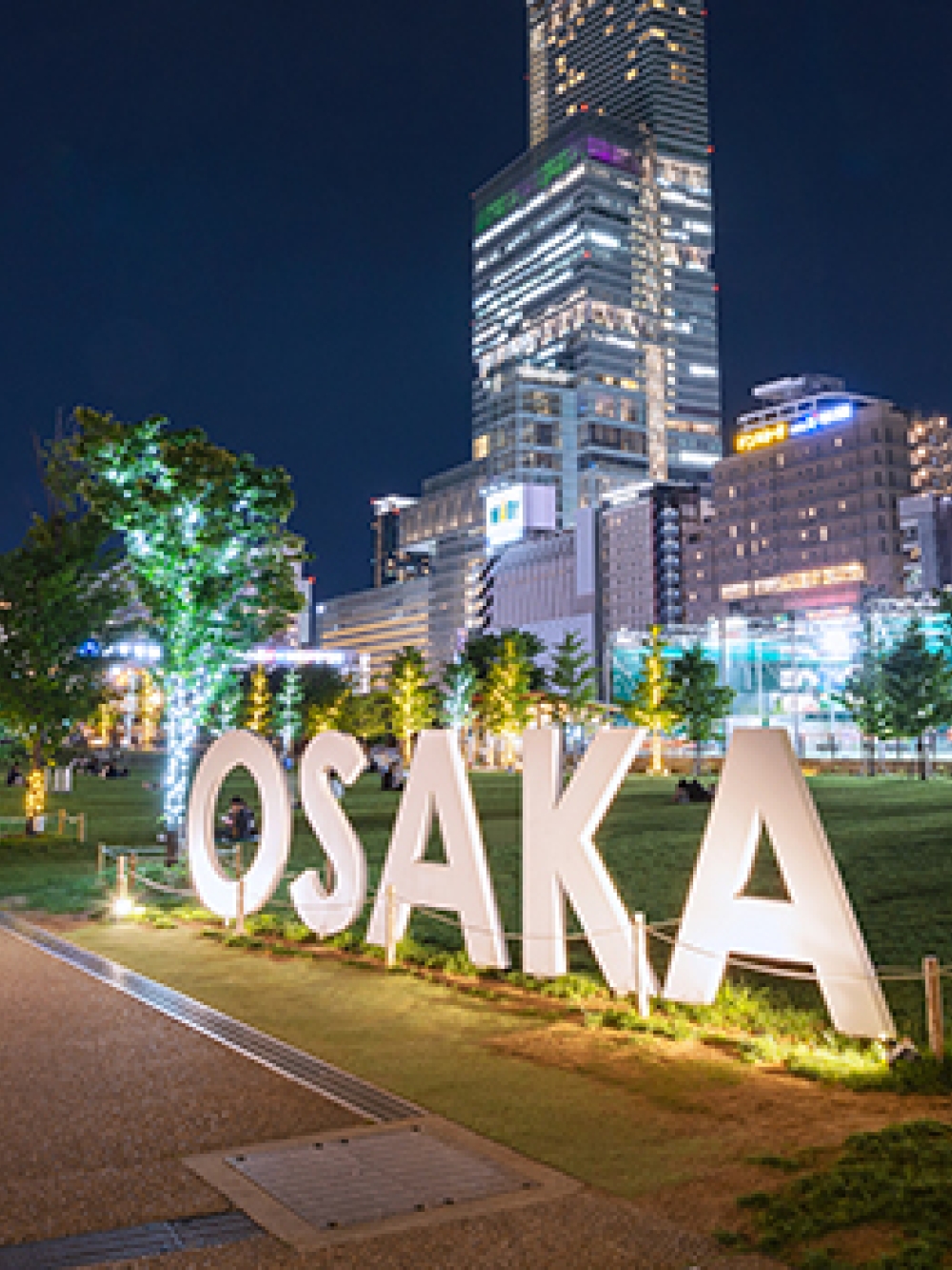 Vue nocturne d’un espace urbain à Osaka, avec de grandes lettres illuminées formant le mot « OSAKA » et des immeubles éclairés en arrière‑plan.