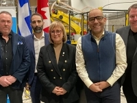 Groupe de cinq personnes posant à l’intérieur d’une usine devant des bannières et des drapeaux, avec de l’équipement industriel en arrière-plan.