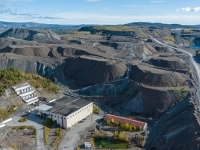 Vue aérienne d’un site minier avec des bâtiments industriels entourés de vastes amas de résidus et de chemins de gravier, dans un paysage vallonné.