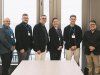 Groupe de personnes debout dans une salle lumineuse avec de grandes fenêtres donnant sur un paysage côtier, posant côte à côte devant une table.